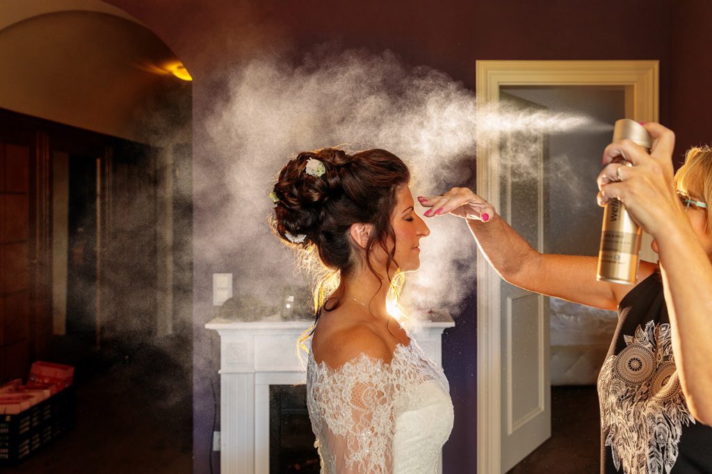 Bei den Vorbereitungen zur Hochzeit auf Schloss Kartzow sprüht die Visagistin fixierendes Spray in die Haare der Braut. Der Hochzeitsfotograf zündet in dem Augenblick den Blitz.