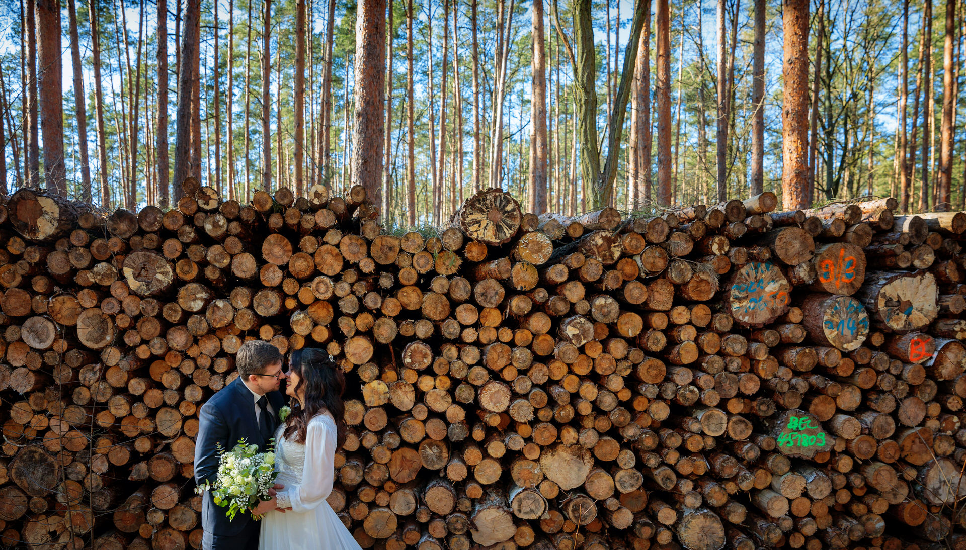 Ein Hochzeitspaar steht innig vor einem großen Stapel gestapelter Holzstämme in einem Wald. Die Braut trägt ein langes, weißes Kleid mit zarten Ärmeln und hält einen Blumenstrauß mit weißen Blüten und grünen Akzenten. Der Bräutigam, in dunklem Anzug und Krawatte, lehnt sich liebevoll zu ihr. Die Holzstämme mit bunten Markierungen und die hohen Kiefern im Hintergrund verleihen der Szenerie eine natürliche, rustikale Atmosphäre.