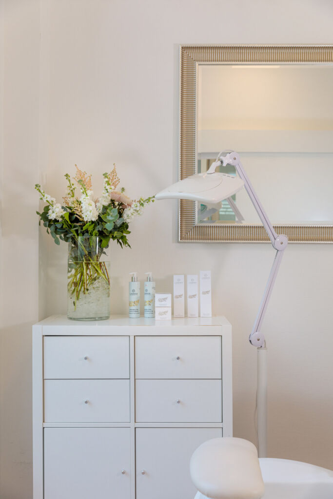 Detailaufnahme eines stilvoll eingerichteten Kosmetikstudios mit einer weißen Kommode, auf der Hautpflegeprodukte und eine Glasvase mit einem eleganten Blumenarrangement stehen. Eine verstellbare Lupenlampe ragt über den Behandlungsbereich, während ein großer Spiegel mit dekorativem Rahmen die helle und moderne Atmosphäre des Raums unterstreicht.