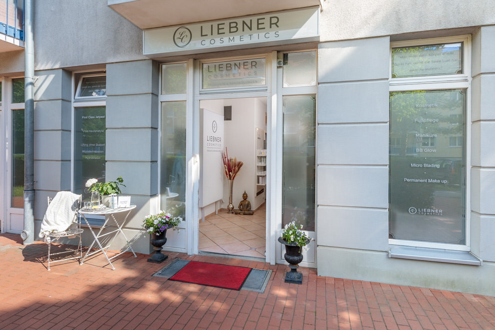 Außenansicht eines Kosmetikstudios mit moderner, heller Fassade und großen Fenstern mit Beschriftung der angebotenen Behandlungen. Der Eingangsbereich ist einladend gestaltet mit einer roten Fußmatte, dekorativen Pflanzen und einem kleinen Tisch mit Stühlen, auf dem Getränke und eine Decke bereitstehen. Die offene Tür gibt einen Blick ins stilvolle Innere des Studios frei.