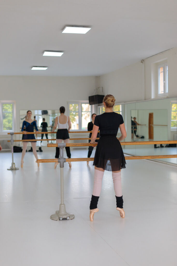 Eine Balletttänzerin steht an der Stange in einem lichtdurchfluteten Tanzstudio und übt konzentriert ihre Haltung. Im Spiegel sind weitere Tänzerinnen zu sehen, die an der Choreografie arbeiten. Dieses Bild zeigt die Eleganz und Disziplin des klassischen Tanzes – ideal für die Social Media Betreuung für Tanzschulen, um die Atmosphäre und Professionalität des Unterrichts authentisch zu vermitteln.