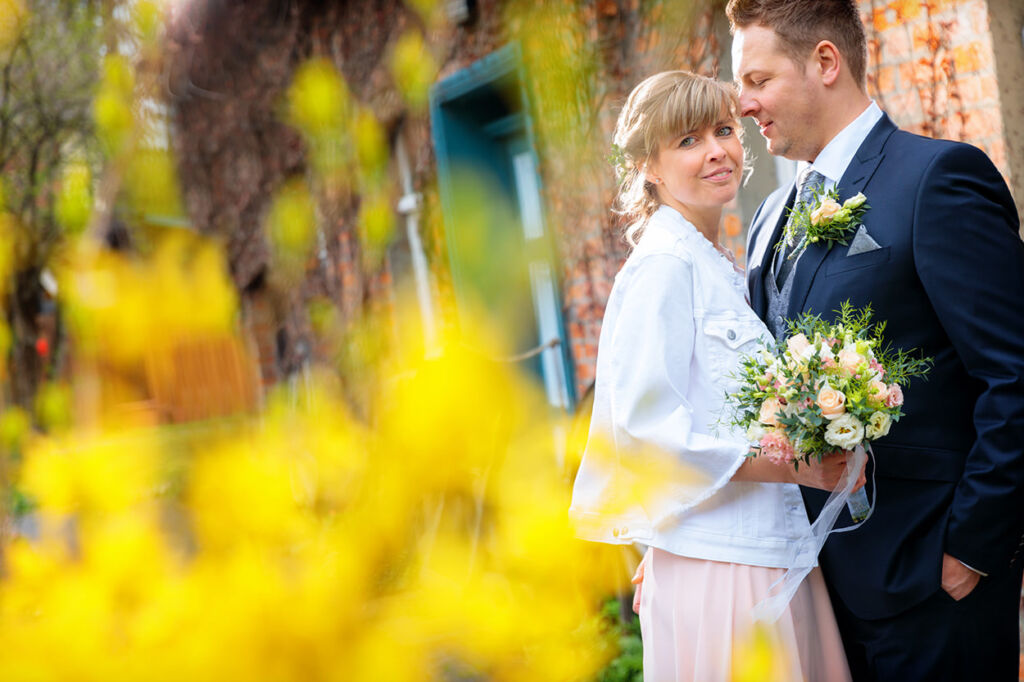 Ein frisch verheiratetes Paar posiert für ein Hochzeitsfoto im Freien. Die Braut trägt eine helle Jacke über ihrem Kleid und hält einen Blumenstrauß mit zarten Pastellfarben. Der Bräutigam trägt einen dunklen Anzug mit einer passenden Blumenanstecknadel. Im Vordergrund sind unscharfe gelbe Blüten zu sehen, die dem Bild eine romantische und warme Atmosphäre verleihen. Im Hintergrund befindet sich eine rustikale Backsteinwand mit einer grünen Tür. Ein frisch verheiratetes Paar posiert für ein Hochzeitsfoto im Freien. Die Braut trägt eine helle Jacke über ihrem Kleid und hält einen Blumenstrauß mit zarten Pastellfarben. Der Bräutigam trägt einen dunklen Anzug mit einer passenden Blumenanstecknadel. Im Vordergrund sind unscharfe gelbe Blüten zu sehen, die dem Bild eine romantische und warme Atmosphäre verleihen. Im Hintergrund befindet sich eine rustikale Backsteinwand mit einer grünen Tür.