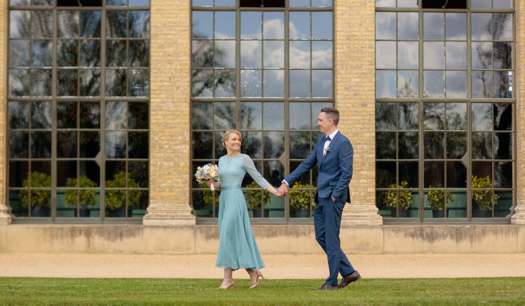 Ein elegantes Paar spaziert Hand in Hand über eine Wiese vor einem historischen Gebäude mit großen Fenstern. Die Frau trägt ein fließendes, pastellblaues Kleid mit Spitzenoberteil und hält einen Blumenstrauß in der Hand. Der Mann trägt einen dunkelblauen Anzug mit passender Ansteckblume. Beide blicken sich lächelnd an, während sie gemeinsam den Moment genießen. Die Reflexionen in den Fenstern und das weiche Frühlingslicht verleihen dem Bild eine romantische Atmosphäre. Ein elegantes Paar spaziert Hand in Hand über eine Wiese vor einem historischen Gebäude mit großen Fenstern. Die Frau trägt ein fließendes, pastellblaues Kleid mit Spitzenoberteil und hält einen Blumenstrauß in der Hand. Der Mann trägt einen dunkelblauen Anzug mit passender Ansteckblume. Beide blicken sich lächelnd an, während sie gemeinsam den Moment genießen. Die Reflexionen in den Fenstern und das weiche Frühlingslicht verleihen dem Bild eine romantische Atmosphäre.