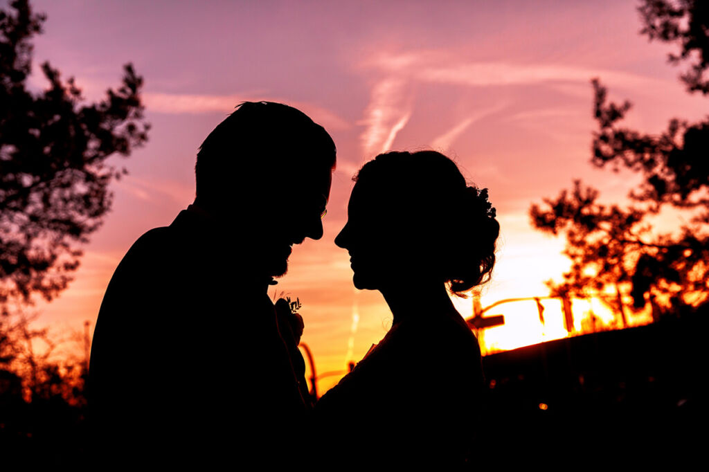 Ein romantisches Silhouettenfoto eines Hochzeitspaares vor einem farbenfrohen Sonnenuntergang sollte immer in euren Zeitplan passen. Die beiden stehen sich nah gegenüber, ihre Gesichter leicht geneigt, während der Himmel in warmen Orange-, Rosa- und Violetttönen leuchtet. Umrahmt von Bäumen entsteht eine stimmungsvolle Atmosphäre, die die emotionale Verbundenheit des Paares unterstreicht. Ein romantisches Silhouettenfoto eines Hochzeitspaares vor einem farbenfrohen Sonnenuntergang sollte immer in euren Zeitplan passen. Die beiden stehen sich nah gegenüber, ihre Gesichter leicht geneigt, während der Himmel in warmen Orange-, Rosa- und Violetttönen leuchtet. Umrahmt von Bäumen entsteht eine stimmungsvolle Atmosphäre, die die emotionale Verbundenheit des Paares unterstreicht.