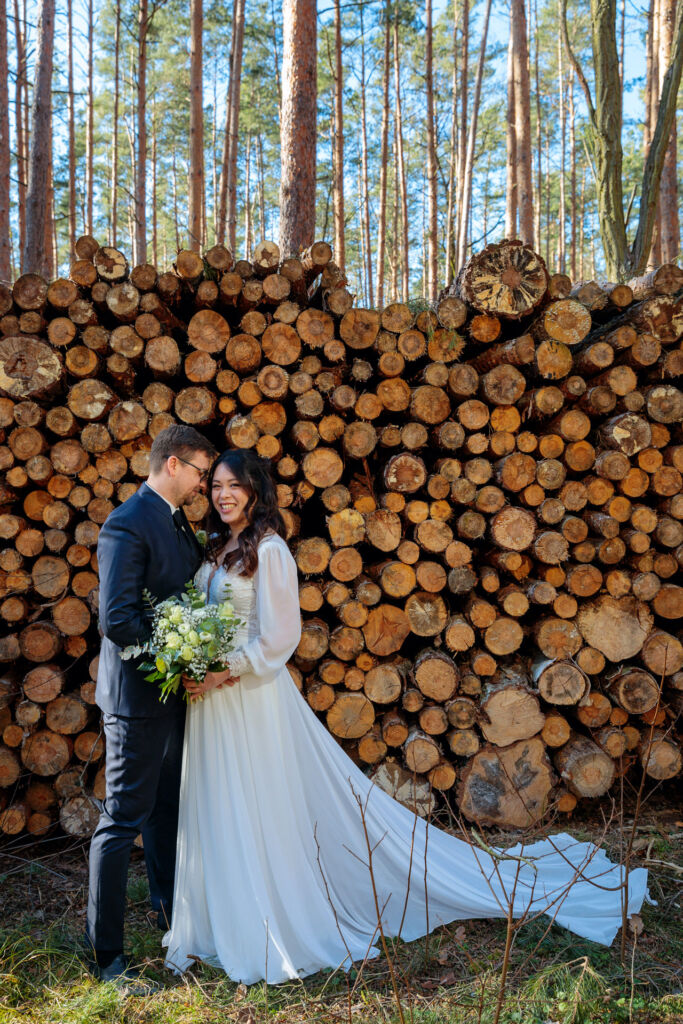 Ein Brautpaar steht lachend vor einem gestapelten Holzstapel im Wald. Die Braut trägt ein fließendes, langärmeliges weißes Hochzeitskleid mit Spitze und eine lange Schleppe, während sie einen natürlichen Brautstrauß mit weißen Blumen und grünen Blättern in den Händen hält. Der Bräutigam, in einem dunklen Anzug mit Weste und Krawatte, lehnt sich liebevoll an sie. Hinter ihnen ragen hohe Kiefern in den blauen Himmel, was der Szenerie eine natürliche und rustikale Atmosphäre verleiht.