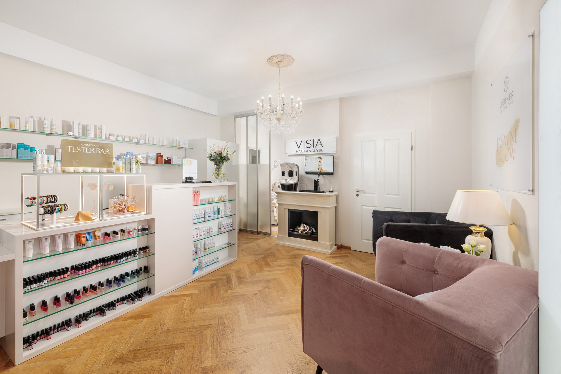 Helles und elegant eingerichtetes Kosmetikstudio mit einer Testerbar für Beauty-Produkte, einem gemütlichen Wartebereich mit samtigen Sesseln und einer modernen Hautanalyse-Station. Die Einrichtung kombiniert weiße Möbel mit Holzboden und einem Kronleuchter, was eine luxuriöse Atmosphäre schafft.