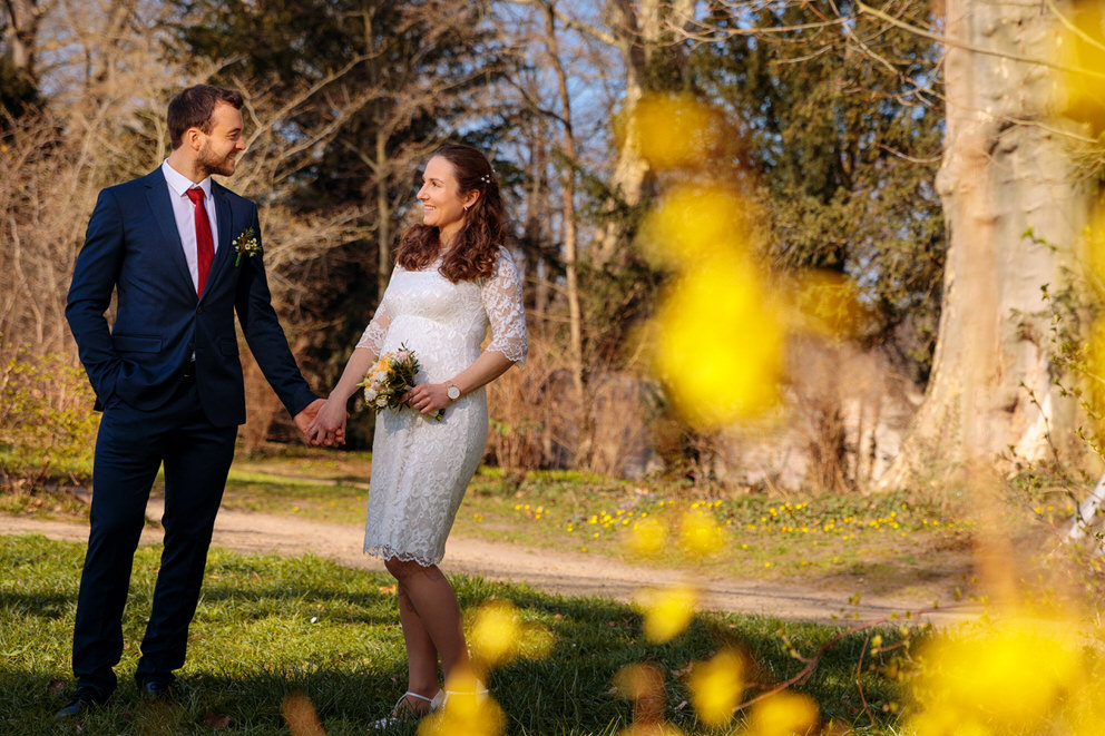 Ein frisch vermähltes Paar steht in einem sonnigen Park und hält sich liebevoll an den Händen. Die Braut trägt ein elegantes, weißes Spitzenkleid mit Dreiviertelärmeln und hält einen kleinen Blumenstrauß. Der Bräutigam trägt einen dunkelblauen Anzug mit roter Krawatte und einem dezenten Anstecker. Im Vordergrund blühen gelbe Frühlingsblüten, während im Hintergrund noch kahle Bäume an den Übergang zwischen Winter und Frühling erinnern. Ein harmonisches Hochzeitsfoto, das die Schönheit einer Frühlingshochzeit einfängt.