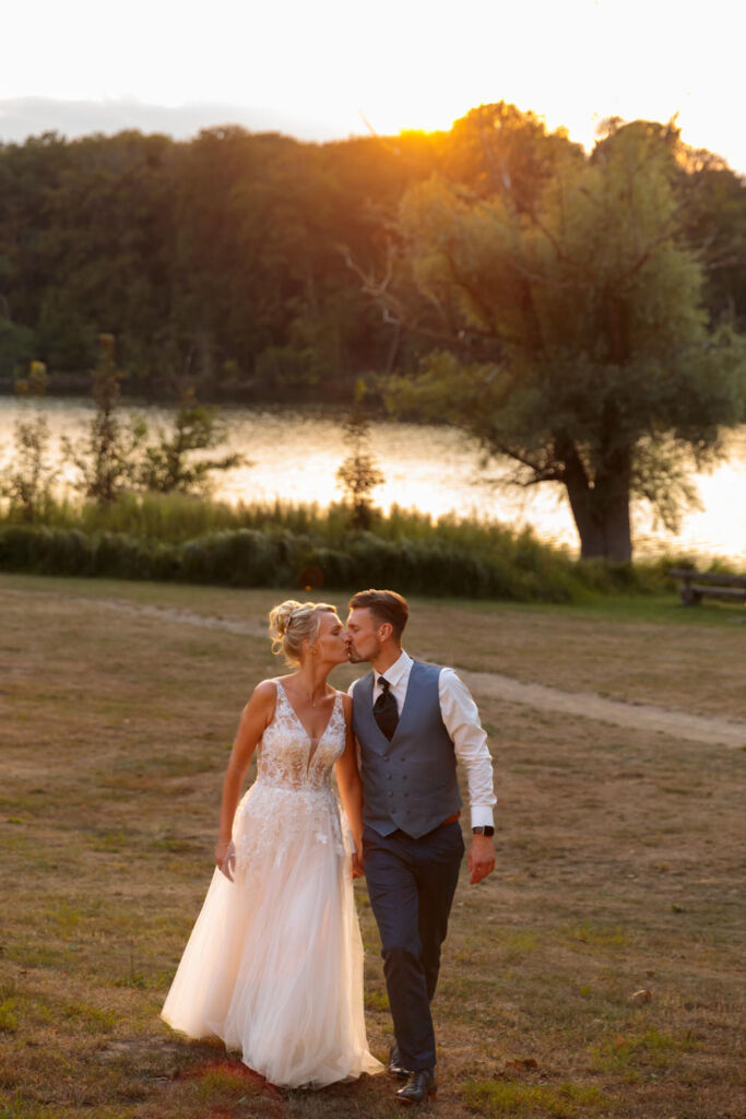 Hochzeitsfotos bei Sonnenuntergang auf dem Landgut Stober Ein Brautpaar küsst sich zärtlich bei Sonnenuntergang am Ufer eines Sees. Die Braut trägt ein elegantes, luftiges Spitzenkleid, der Bräutigam eine Weste mit Krawatte und weißem Hemd.