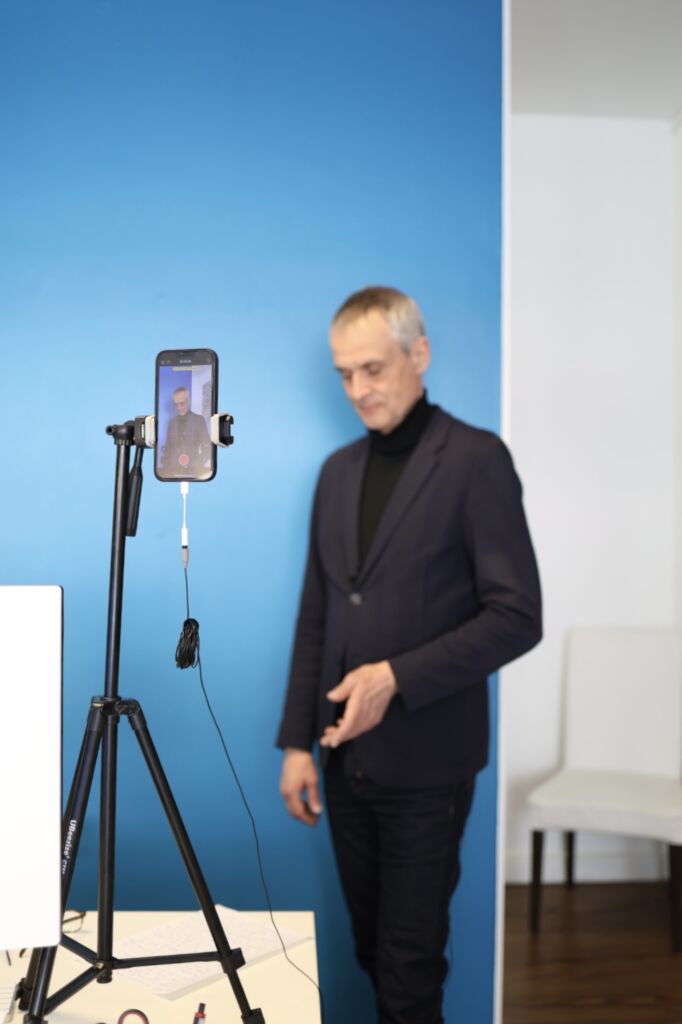 Ein Immobilienmakler steht vor einer blauen Wand und nimmt mit seinem Smartphone ein Video auf, das auf einem Stativ befestigt ist. Das Setup mit Mikrofon und Licht zeigt einen professionellen Ansatz für digitale Kommunikation. 