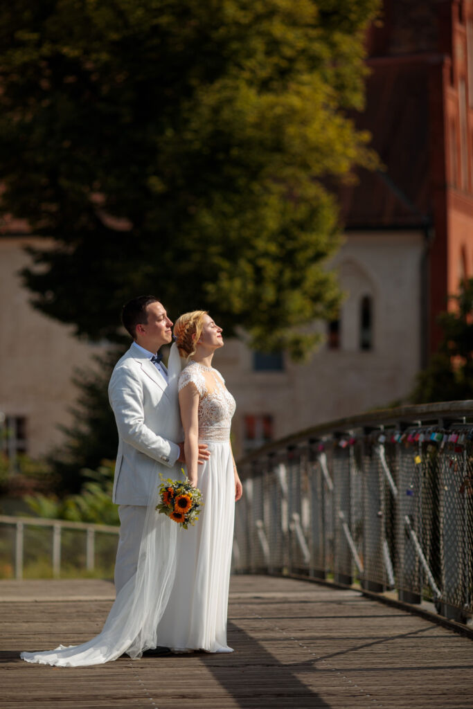 Ein Hochzeitspaar steht auf einer romantischen Brücke mit zahlreichen Liebesschlössern, während sie gemeinsam die warmen Sonnenstrahlen genießen.