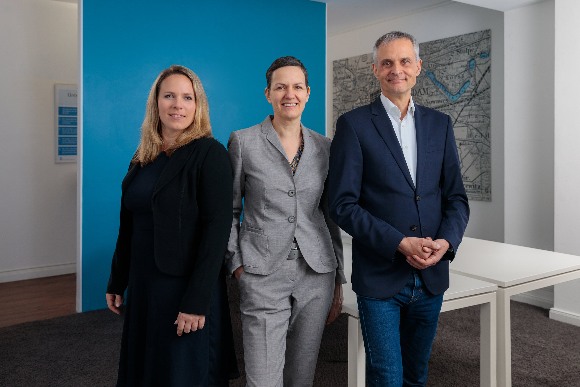 Drei Immobilienmakler stehen in einem modern eingerichteten Büro vor einer blauen Wand und lächeln professionell in die Kamera. Das Team vermittelt Kompetenz und Vertrauen – essenzielle Werte in der Immobilienbranche. Dieses Bild eignet sich ideal für die Social Media Betreuung für Immobilienmakler, um die persönliche und fachliche Expertise ansprechend zu präsentieren und potenzielle Kunden anzusprechen.
