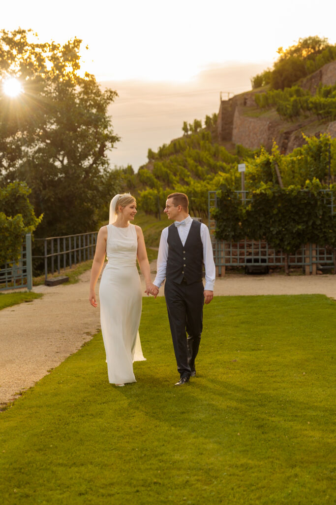 Ein glückliches Hochzeitspaar spaziert Hand in Hand durch eine idyllische Weinberg-Landschaft bei Sonnenuntergang.