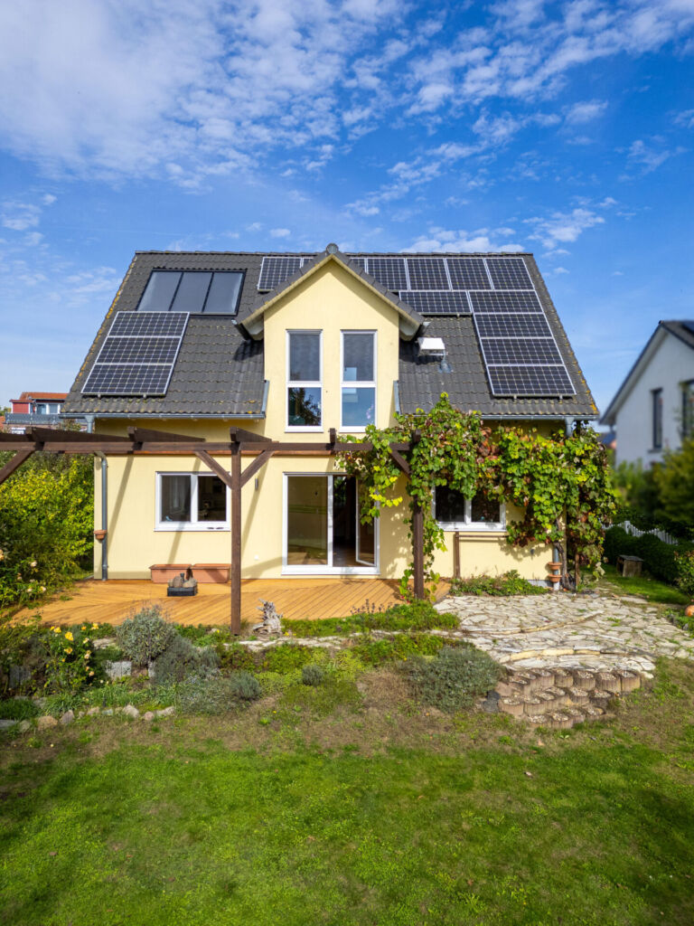 Ein modernes Einfamilienhaus mit gelber Fassade, großer Terrasse und üppig bewachsener Pergola steht im Grünen. Auf dem Dach sind Solarpanele installiert, was auf eine nachhaltige Energieversorgung hinweist.