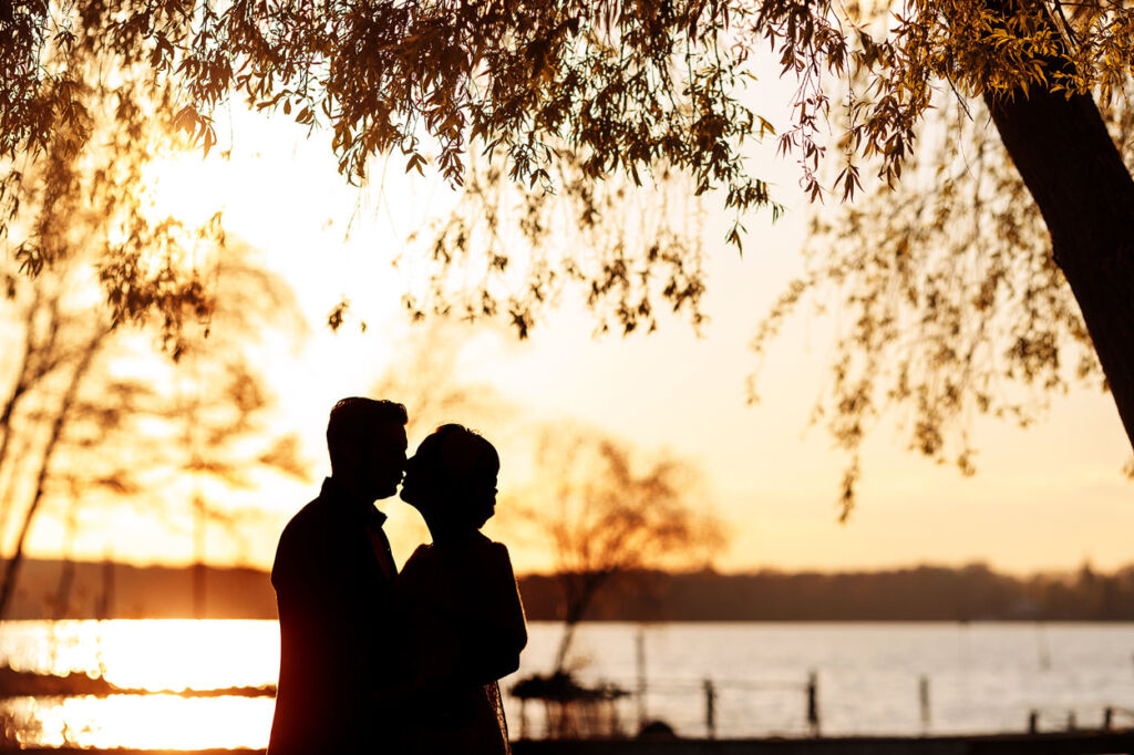 Ein verliebtes Paar steht in inniger Umarmung am Ufer eines Sees und genießt den Moment bei Sonnenuntergang. Die Silhouetten der beiden heben sich romantisch vor dem goldenen Licht des Abends ab, während herabhängende Äste den oberen Bildrand umrahmen. Die warme Atmosphäre und die Spiegelung des Lichts im Wasser machen dieses Bild zu einer perfekten Hochzeits- oder Paarfotografie mit magischer Stimmung.