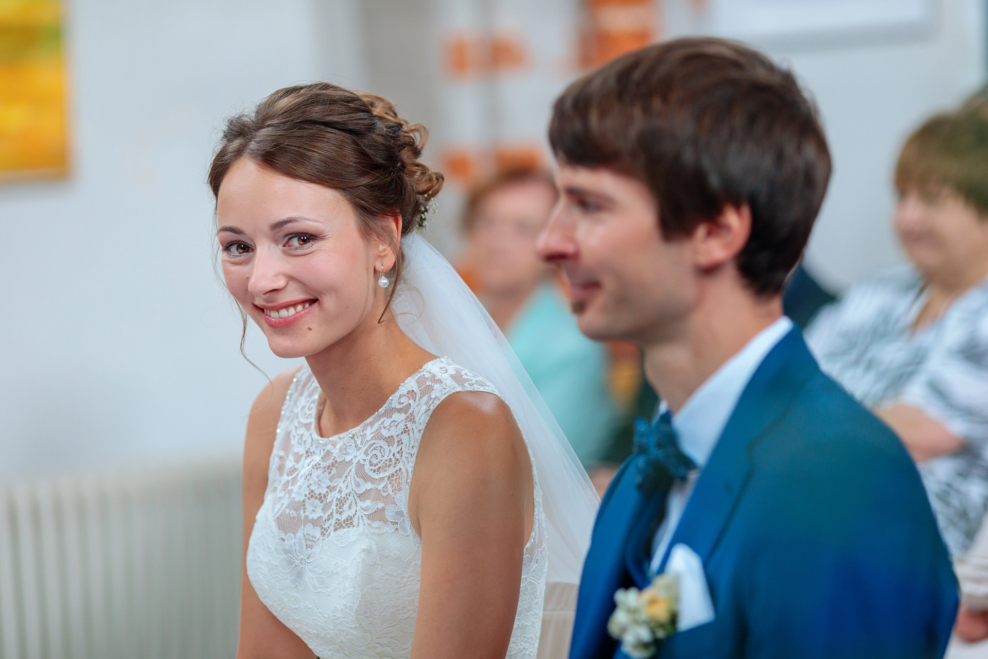 Nahaufnahme einer lächelnden Braut während der Trauung in einer Kirche. Sie trägt ein elegantes Spitzenkleid mit Schleier und schaut glücklich in die Kamera, während der Bräutigam in einem blauen Anzug unscharf im Vordergrund sitzt. Im Hintergrund sind unscharf Gäste der Zeremonie zu sehen. Ein Moment voller Vorfreude und Emotion, festgehalten von den bei vielen Hochzeiten zu zweit arbeitenden Hochzeitsfotografen Michael Reinhardt und Christian Sommer