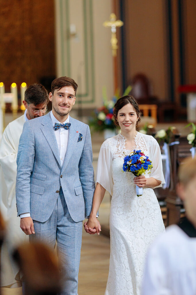Ein frisch vermähltes Paar verlässt Hand in Hand die Kirche nach der Trauung. Die Braut trägt ein elegantes, cremefarbenes Spitzenkleid und hält einen farbenfrohen Brautstrauß in Blau und Gelb. Der Bräutigam trägt einen hellblauen Anzug mit passender Fliege. Beide blicken glücklich in die Kamera – ein emotionaler Moment direkt nach dem Ja-Wort.